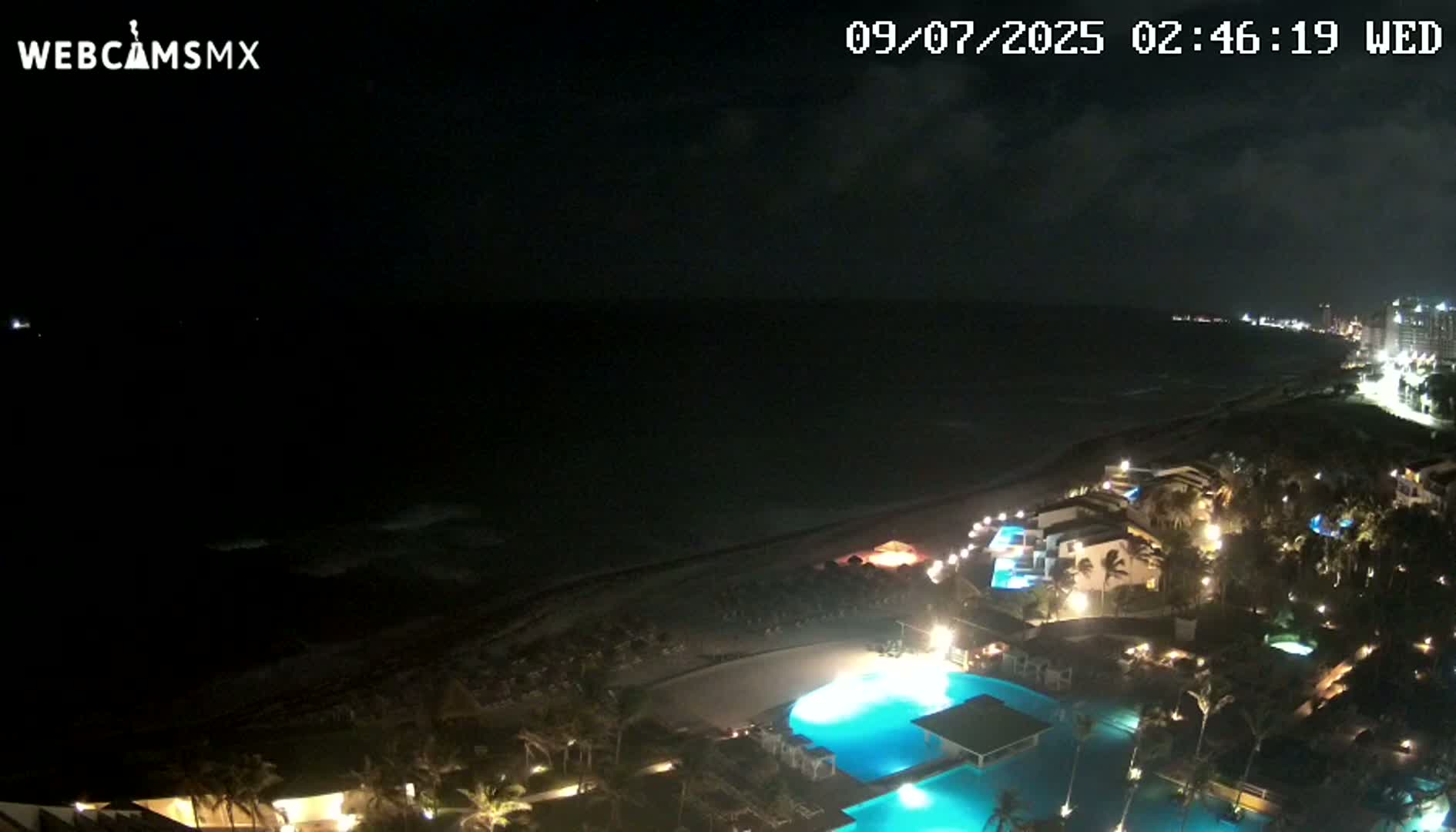 A nighttime view of a resort area along a dark ocean under a cloudy sky shows illuminated pools and buildings, a beach, and a distant shoreline.