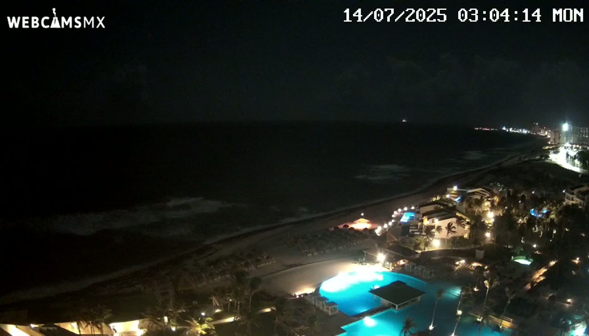A nighttime view shows a dark ocean with waves lapping a sandy beach, next to a resort with illuminated pools and buildings.