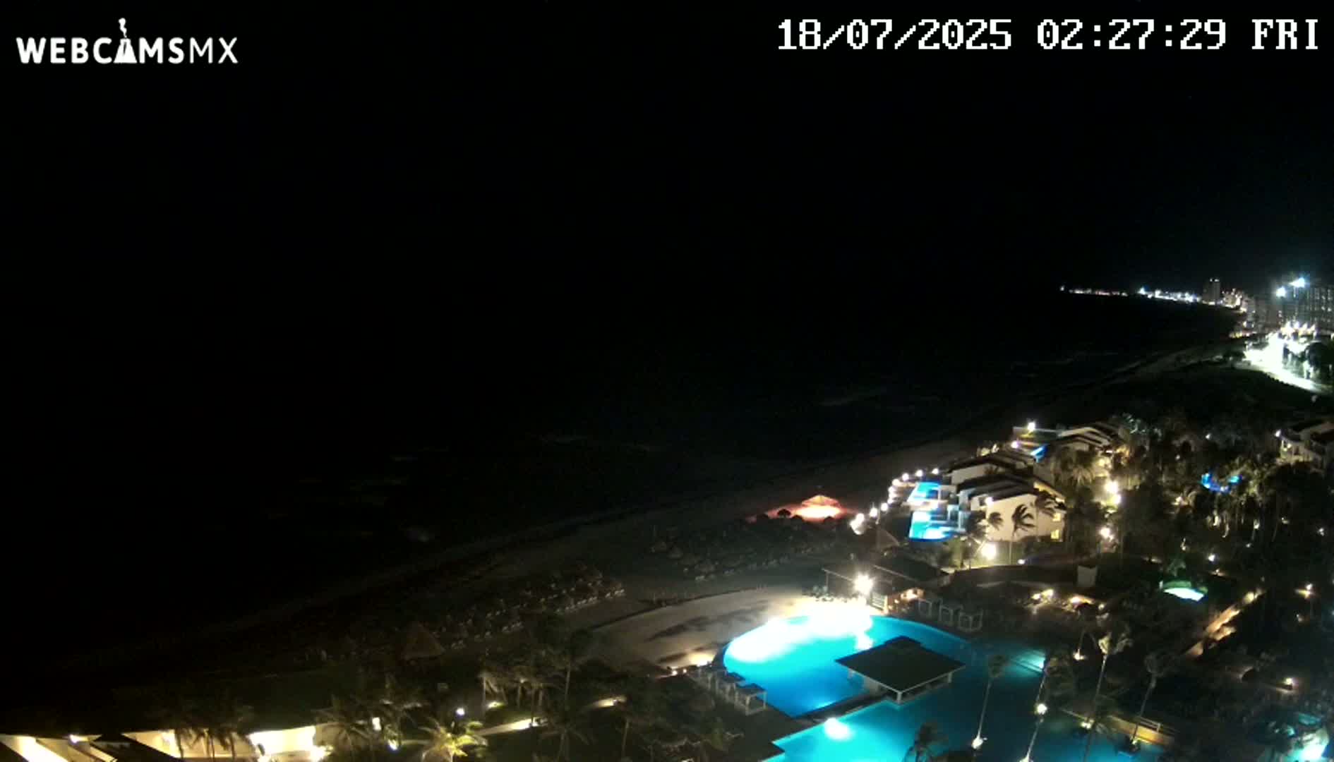 A nighttime aerial view shows a resort complex with illuminated pools and buildings along a dark ocean beach under a clear, dark sky.