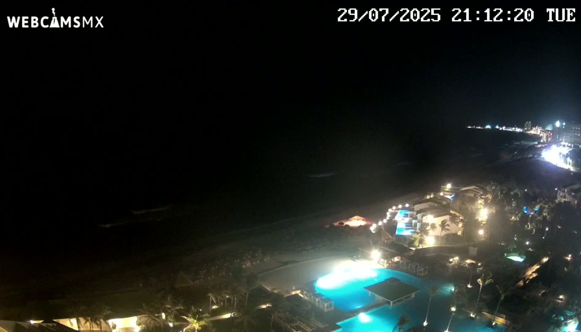 A nighttime aerial view shows a resort complex with illuminated pools and buildings along a dark ocean beach under a clear, dark sky.
