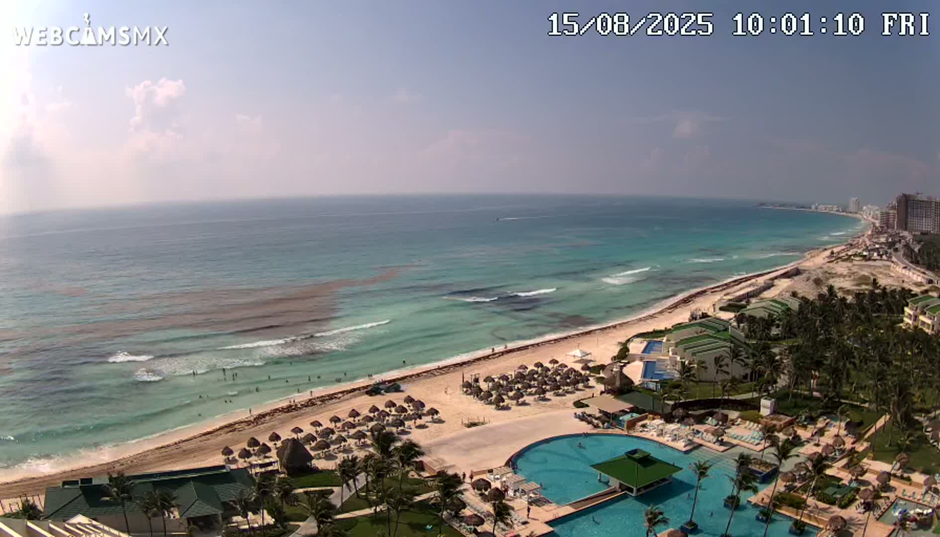 An aerial view shows a beach resort on a sandy coast with a turquoise ocean under a mostly sunny sky, featuring moderate waves and some seaweed along the shore.