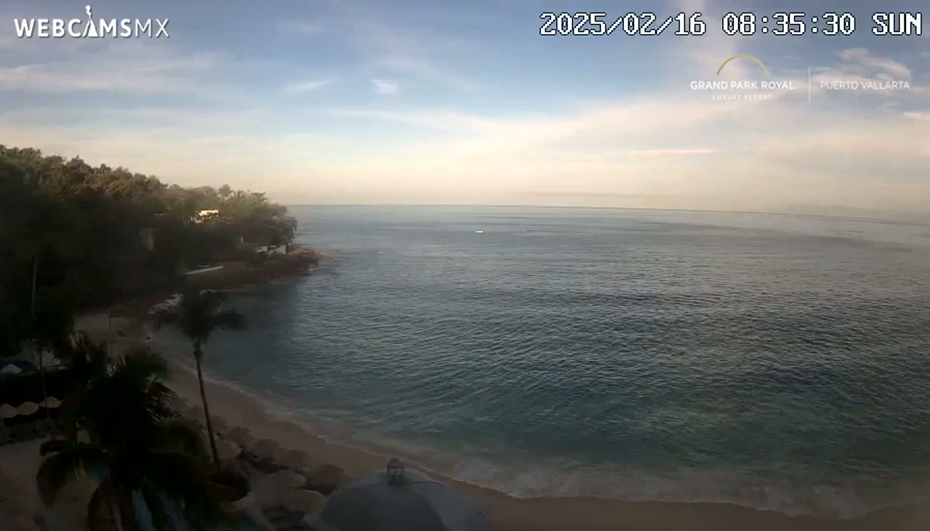 A partly sunny morning shows a secluded cove with a sandy beach and calm ocean water, bordered by lush green vegetation.