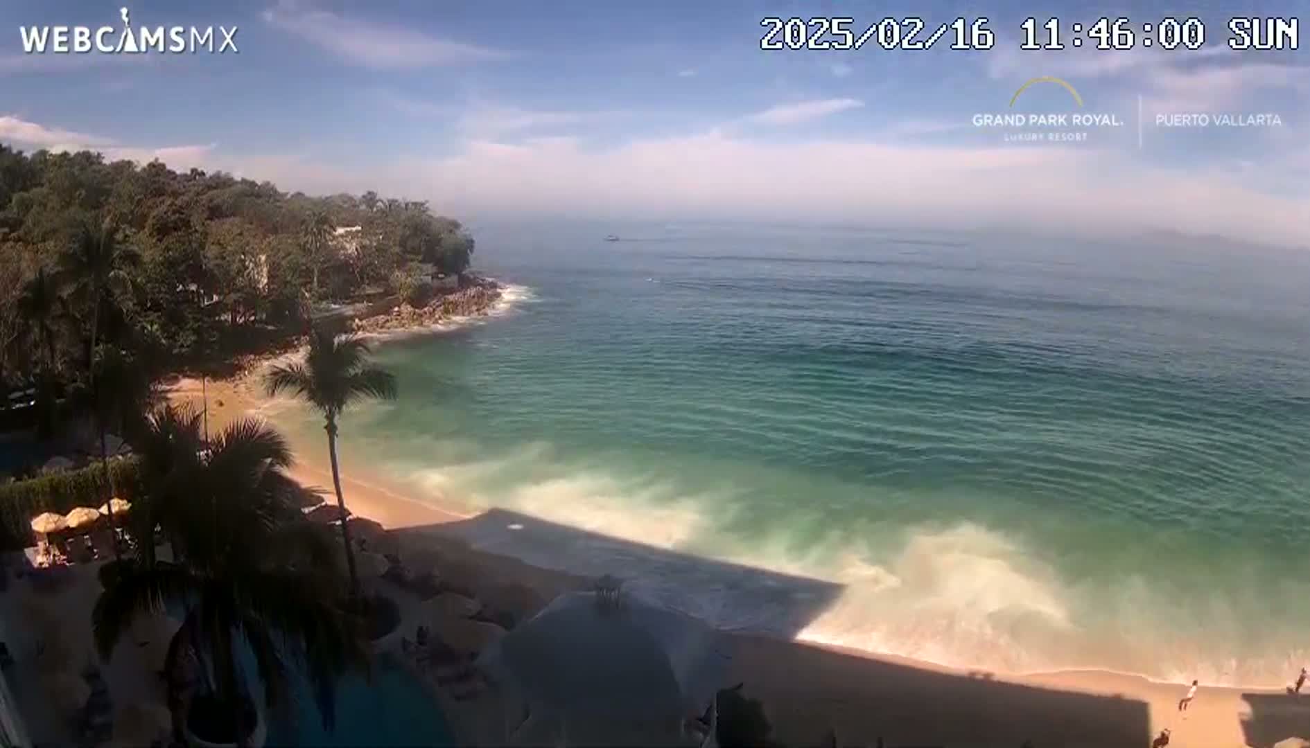 A sunny day reveals a tranquil beach scene with turquoise water lapping gently on a sandy shore, backed by lush green vegetation and a resort area.
