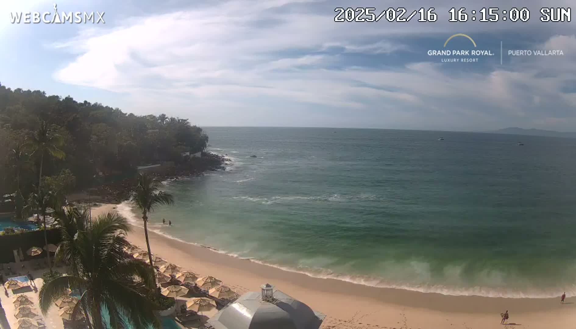 A partly sunny day at a beach with calm waves, showing a sandy shore, palm trees, and resort umbrellas.