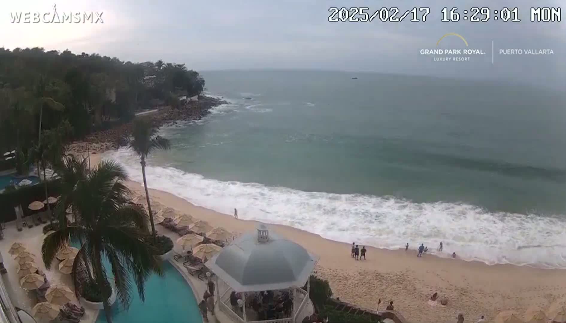 An overcast day at a beach resort shows a sandy beach with moderate waves, a swimming pool, and palm trees.