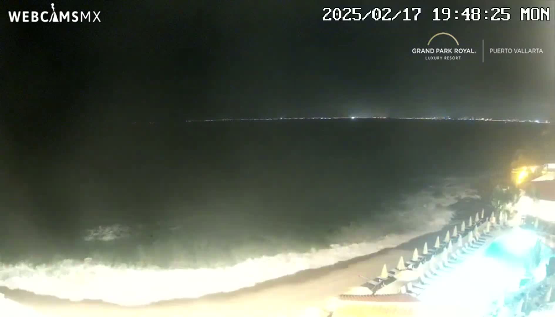 A nighttime beach scene shows waves lapping onto the shore near a resort with beach chairs and a lit pool.