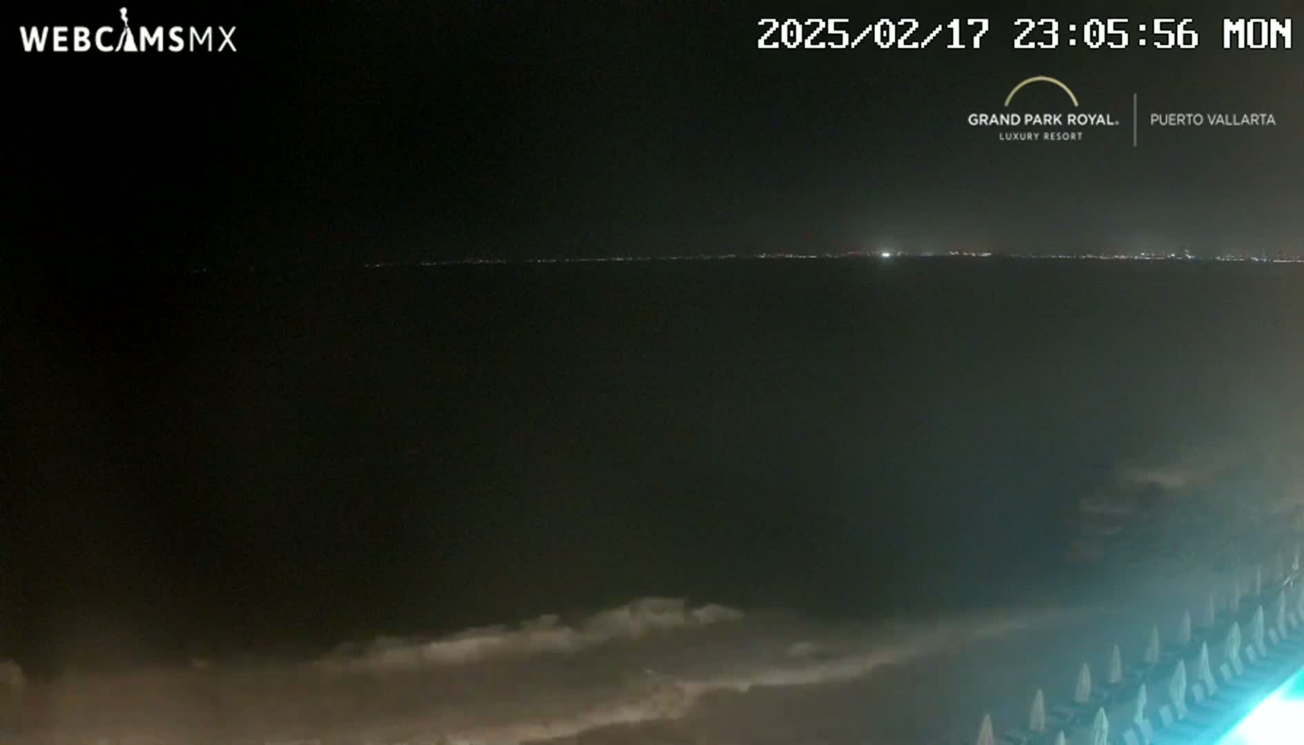 A nighttime coastal scene shows a dark ocean with a distant line of city lights, and in the foreground, a beach with several rows of lounge chairs.