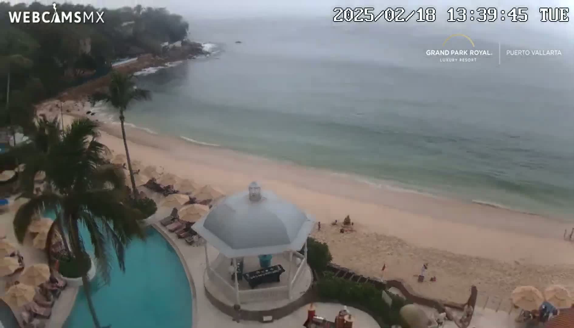 An aerial view shows a beach with light-colored sand, a calm ocean, a resort pool and gazebo, palm trees, and overcast weather.