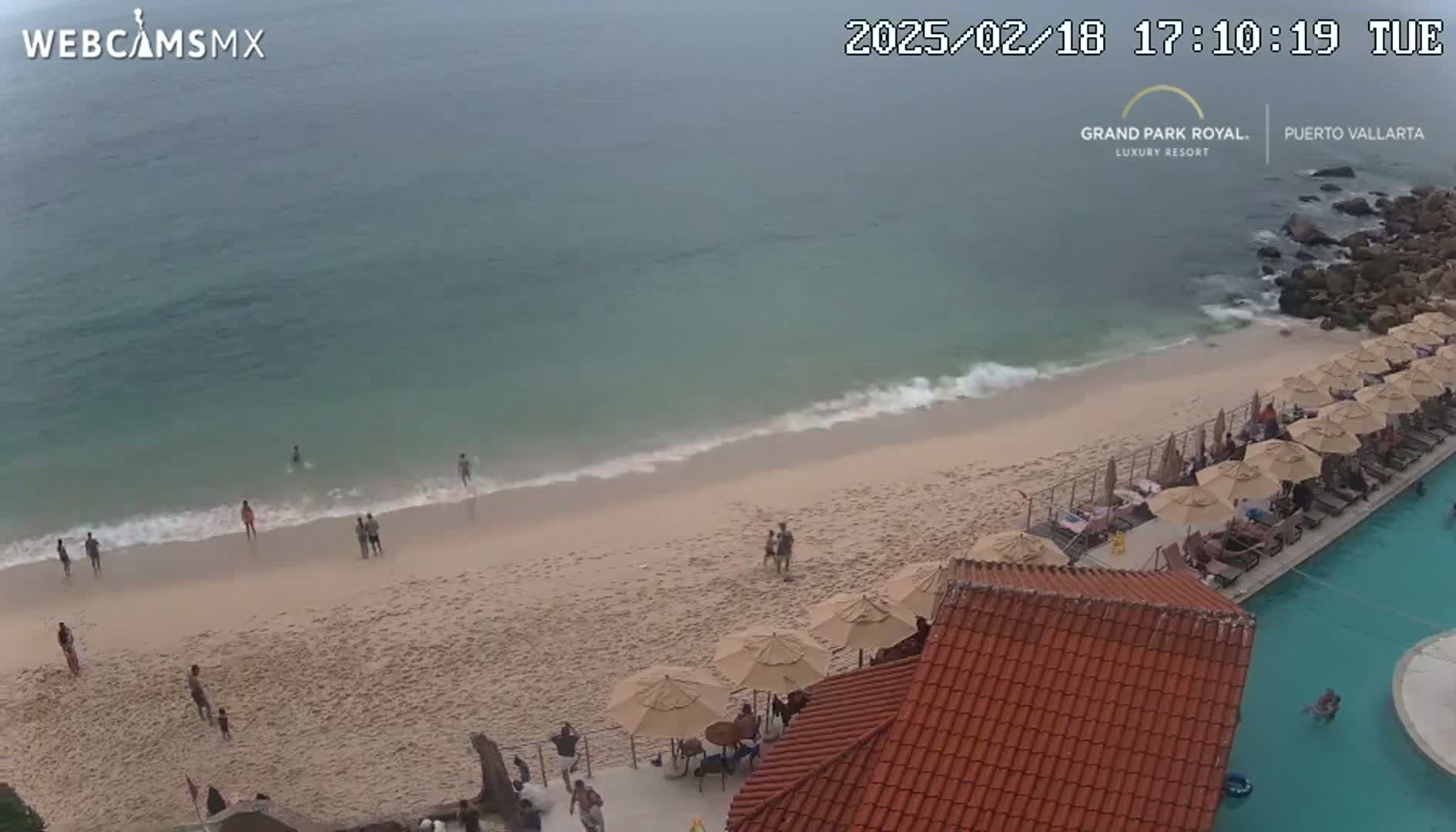 An aerial view shows a sandy beach with a calm, light-green ocean, several people swimming and walking on the beach, and a resort pool and patio area with numerous umbrellas.