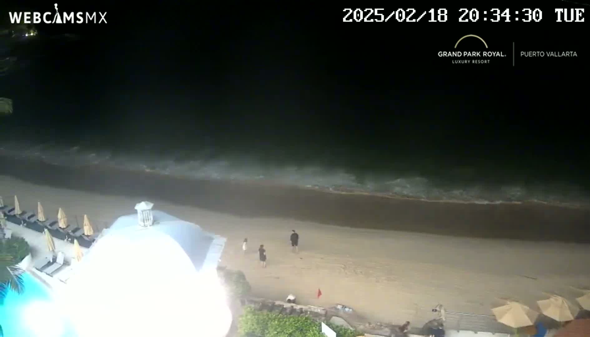 At night, a sandy beach with gentle waves laps the shore near a resort with a pool and several beach umbrellas.