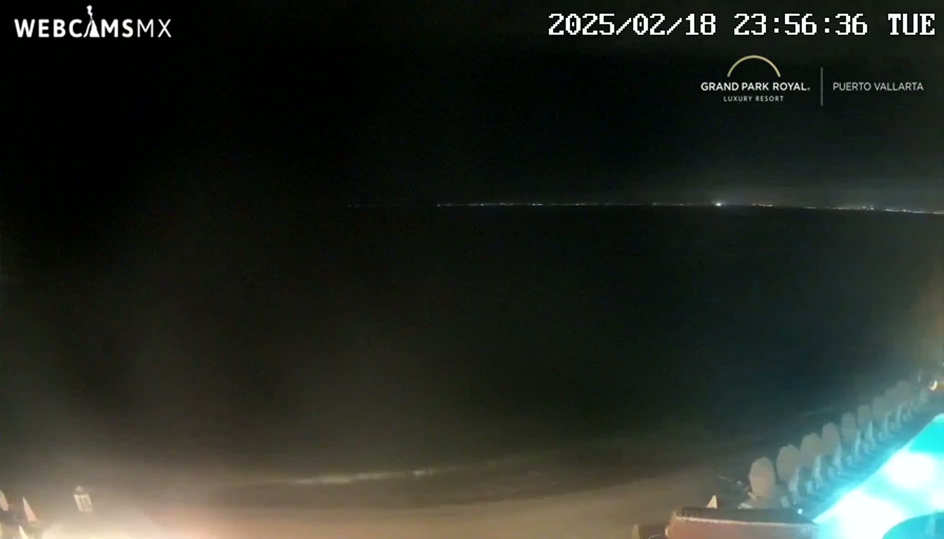 A nighttime view of a dark ocean with a faint, distant horizon light, and a pool area with illuminated chairs in the foreground.