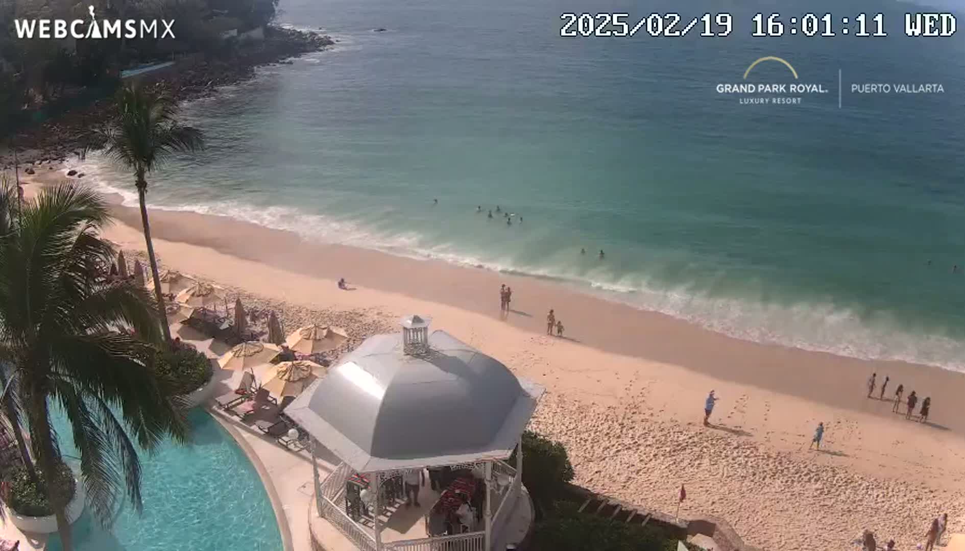 An aerial view shows a sandy beach with gentle waves lapping the shore, a few people swimming and sunbathing, palm trees, a pool, and a gazebo near a resort.
