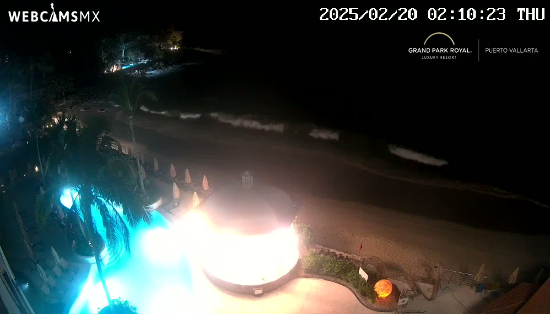 A nighttime view of a resort pool lit in bright blue, next to a beach with gentle waves under a dark sky.