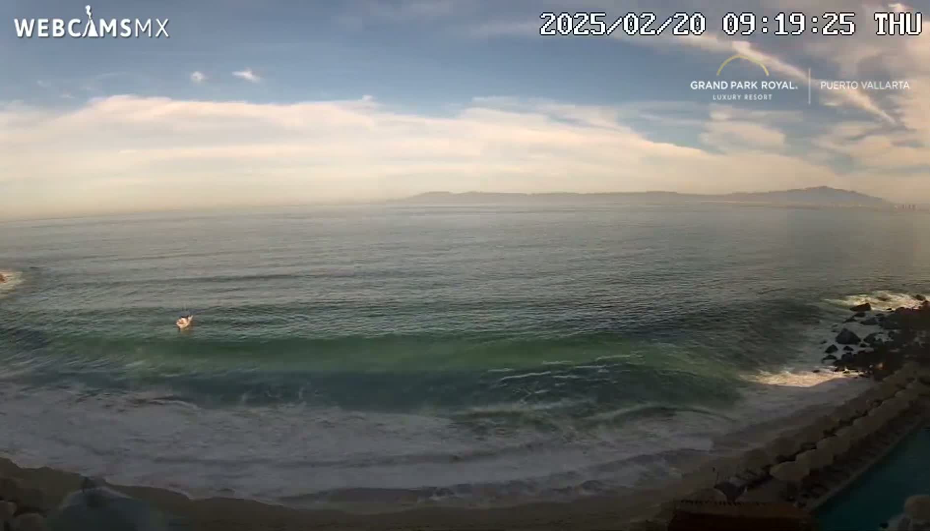A partly cloudy sky overlooks a calm ocean meeting a sandy beach with small waves, and a distant mountain range.