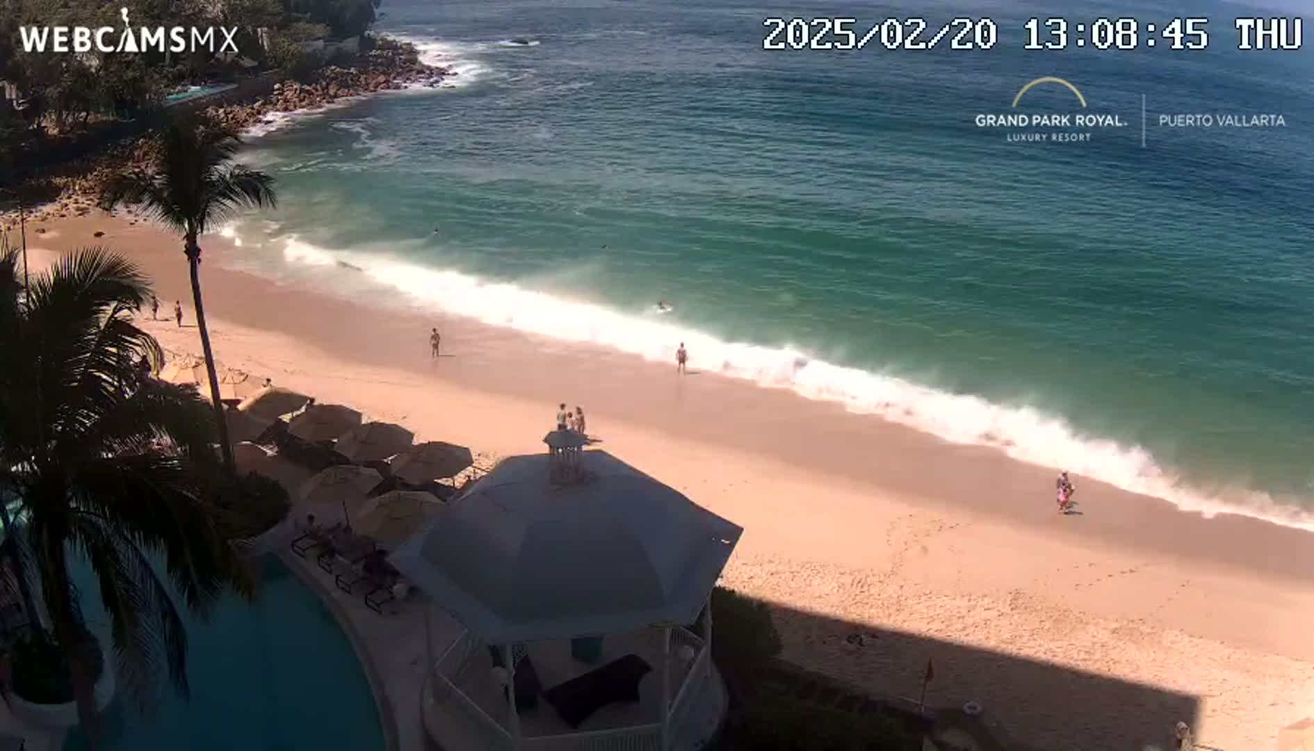 An aerial view shows a sandy beach with a few people on it, gentle waves lapping the shore, and a partially shaded gazebo near a swimming pool; the weather is sunny.
