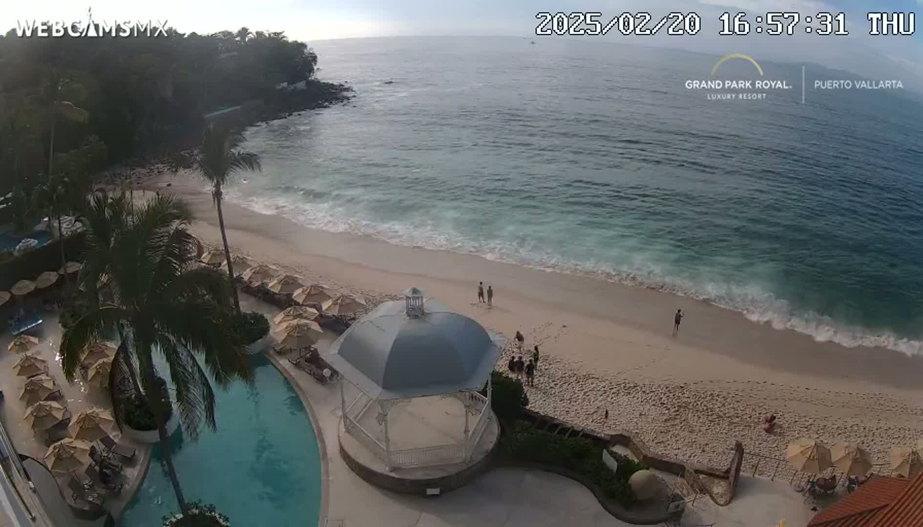 An aerial view shows a sandy beach with gentle waves lapping the shore, a resort pool, and a gazebo, all under a mostly clear sky.