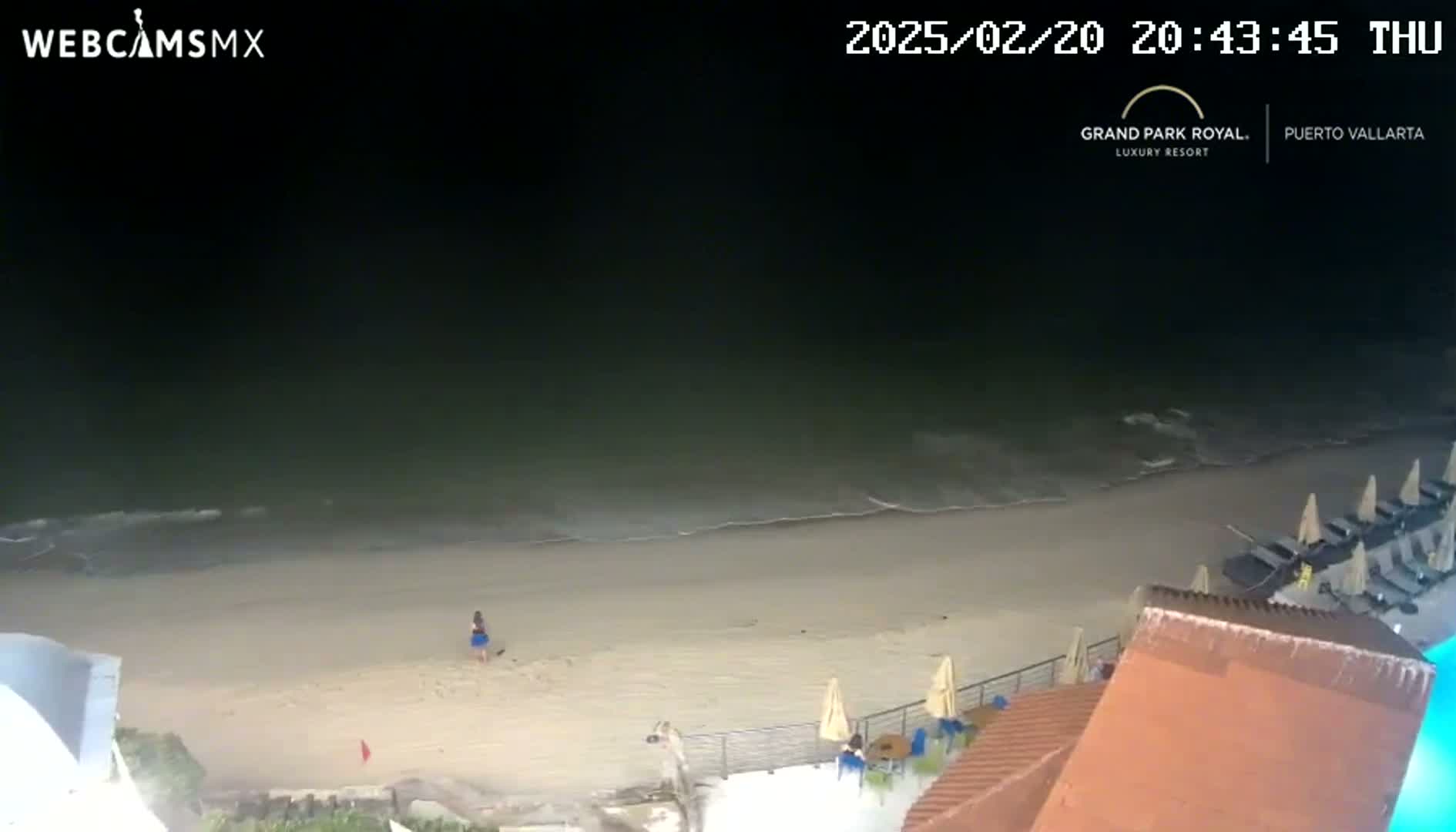 A nighttime beach scene shows calm waves lapping a sandy shore, with a few people visible near some beach umbrellas and lounge chairs near a building.