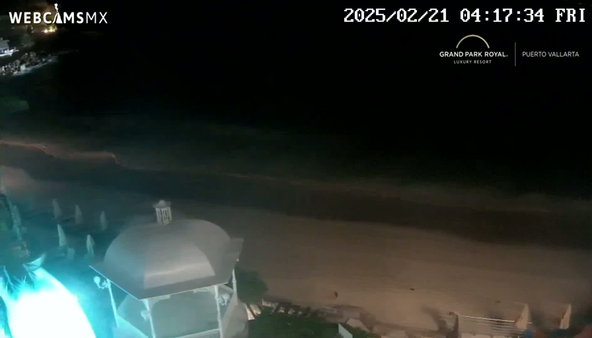 A nighttime view of a beach with calm water, a sandy shore, and a partially visible gazebo illuminated in teal.