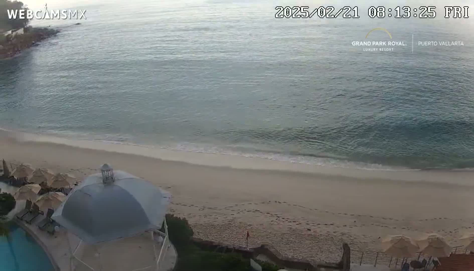 A sandy beach with calm, light-blue water and a few footprints on the sand is overlooked by a gazebo and closed umbrellas near a swimming pool.