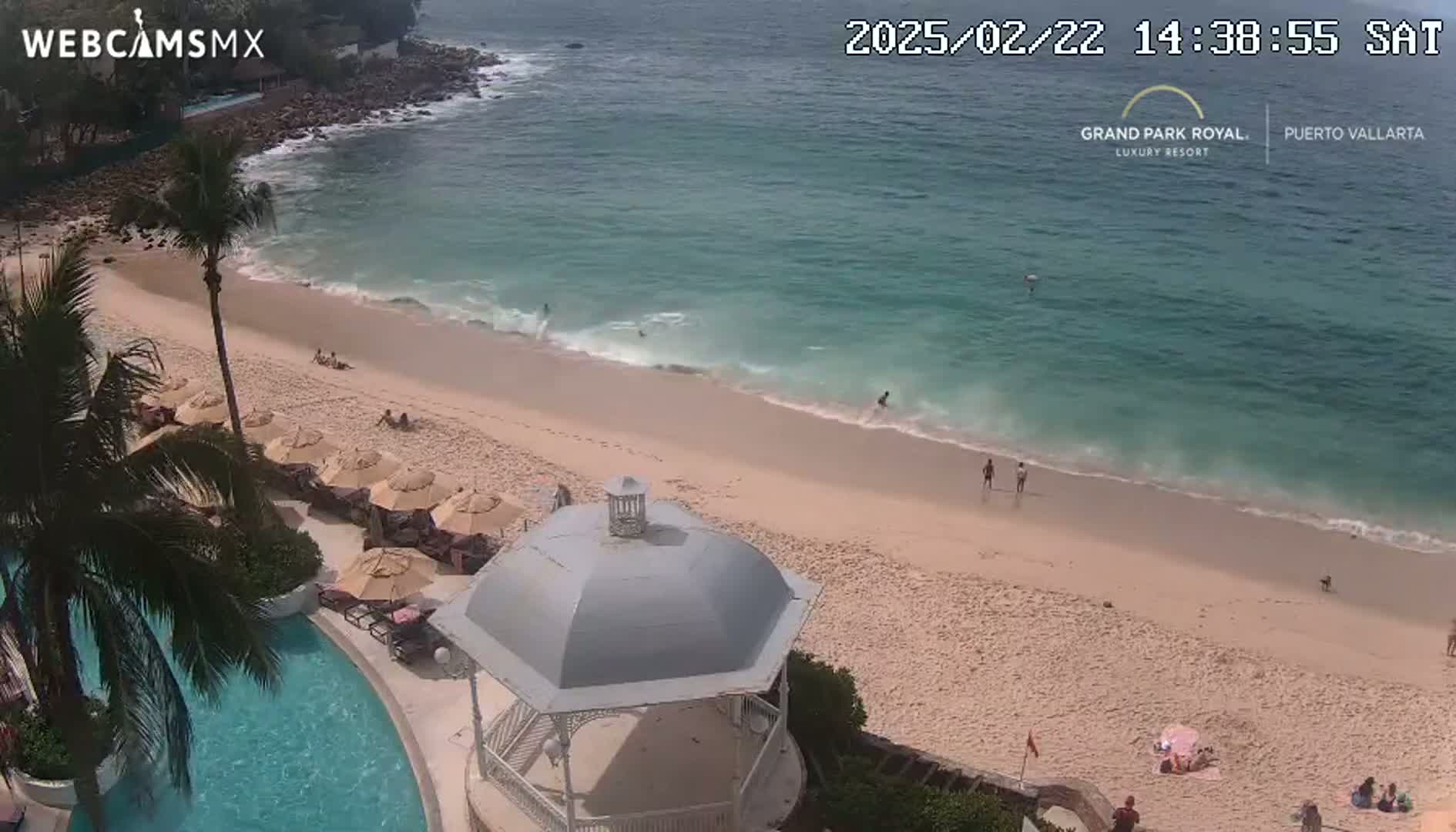 An aerial view shows a sandy beach with calm, clear turquoise water, several people enjoying the beach, and a gazebo near a resort pool under a sunny sky.