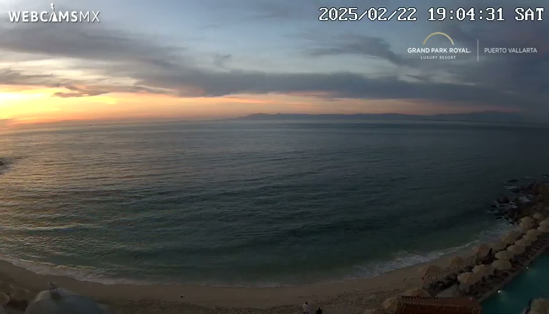 A tranquil ocean sunset scene shows a sandy beach with beach umbrellas and a calm sea under a partly cloudy sky.