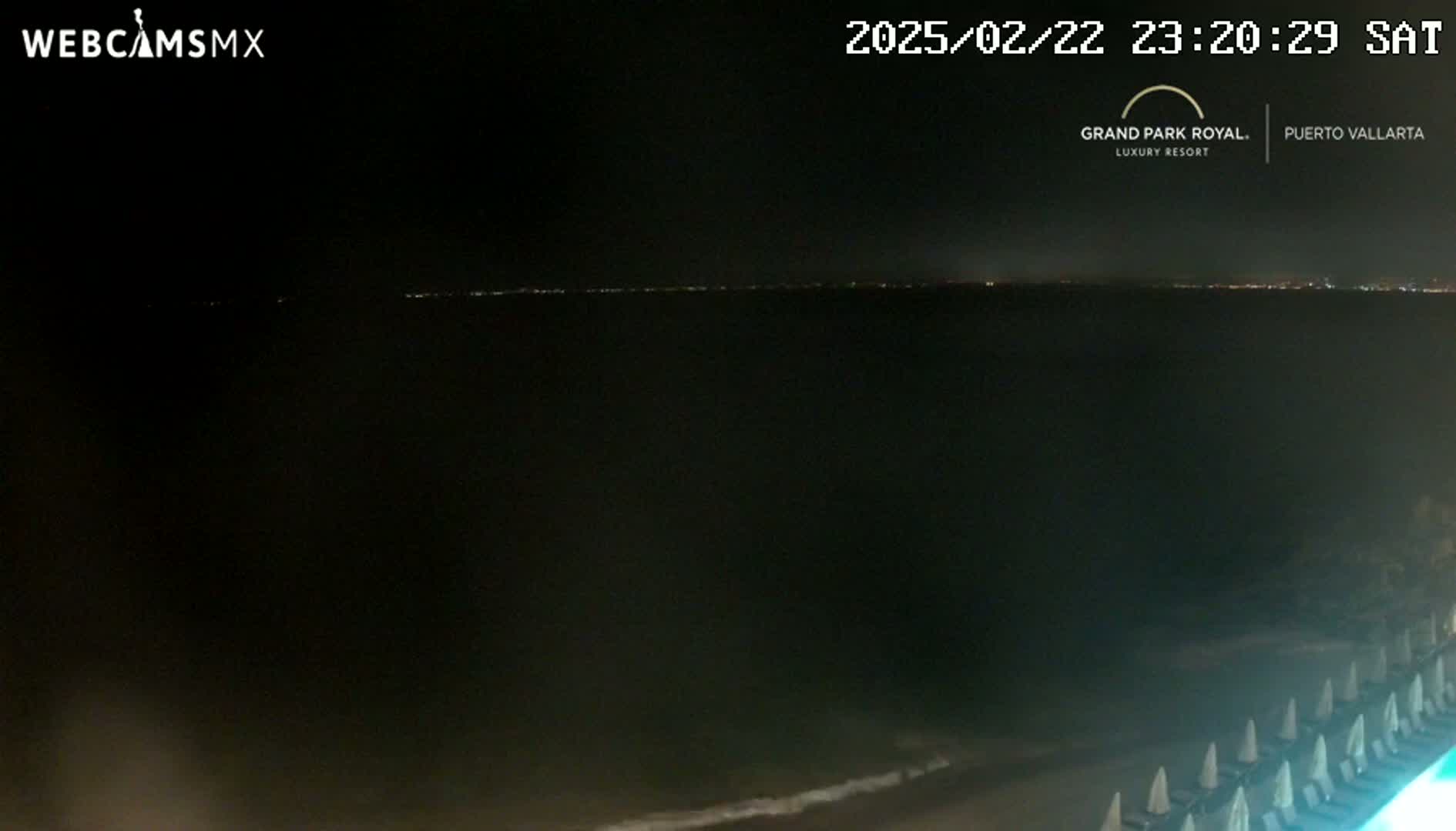 A nighttime view of a dark ocean with a distant city skyline illuminated, a beach with lounge chairs, and calm waves.