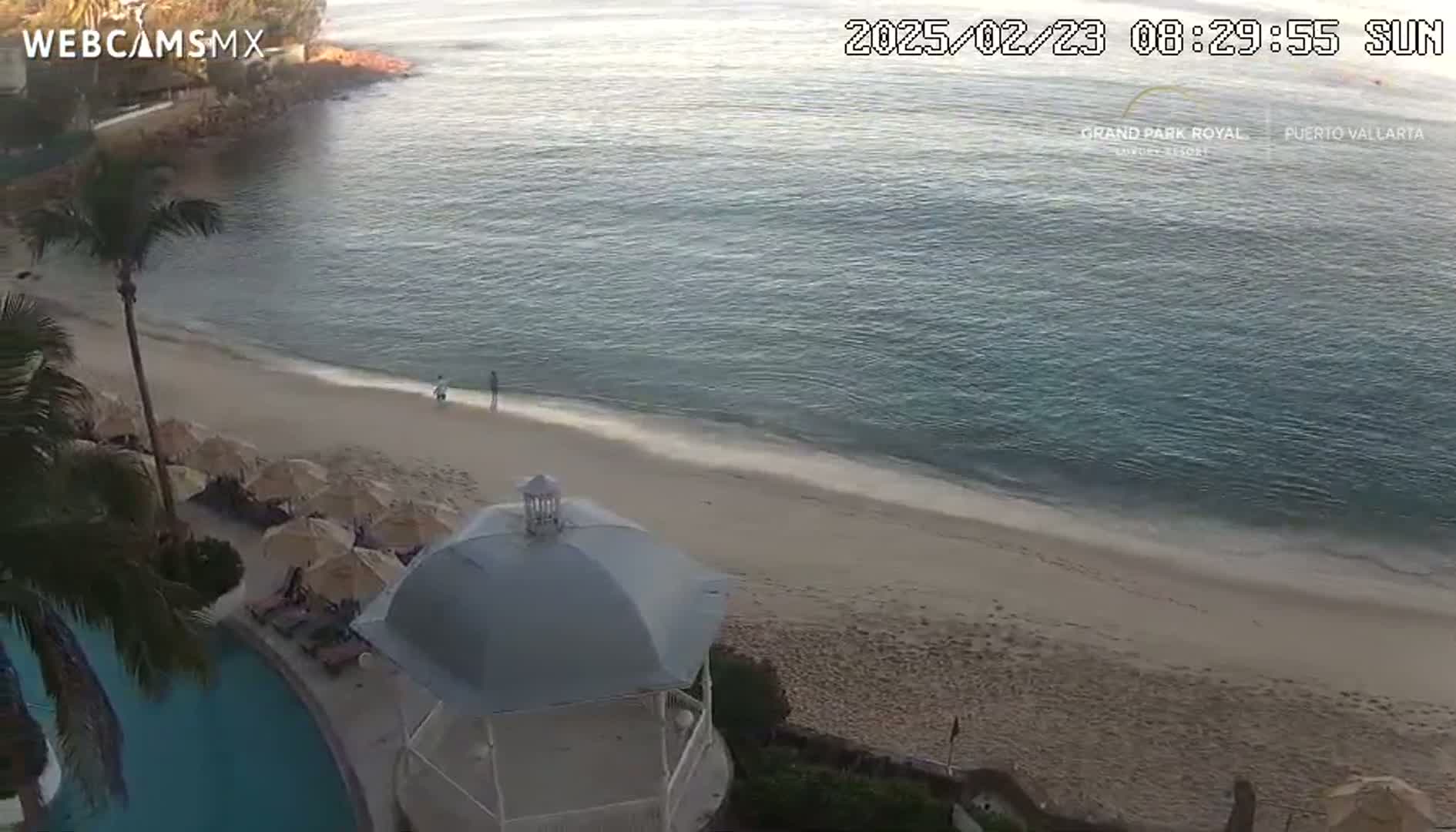 A sandy beach with calm, clear water stretches out from a resort area with a swimming pool and a gazebo under a sunny sky.