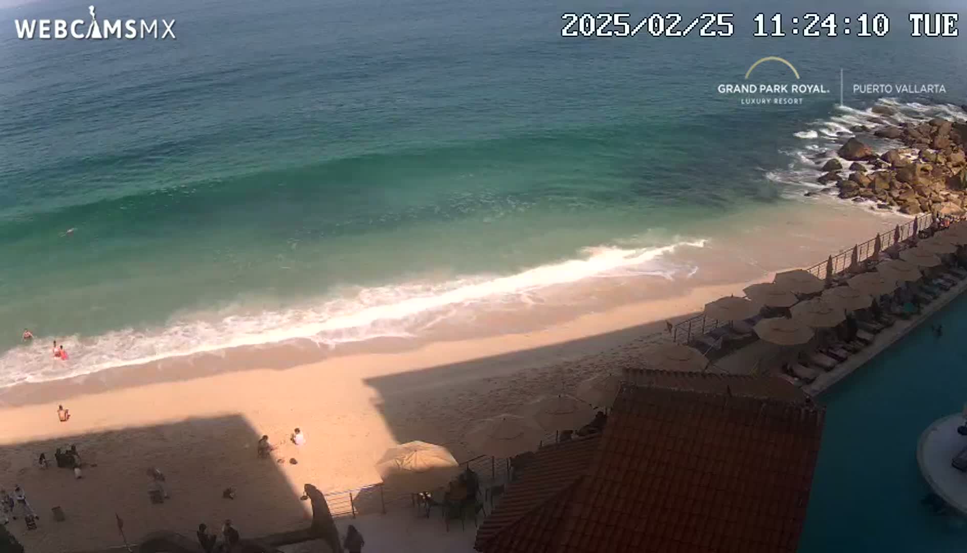 An overhead view shows a sandy beach with gentle waves lapping the shore, several people on the sand, and a resort pool and patio area with numerous umbrellas.