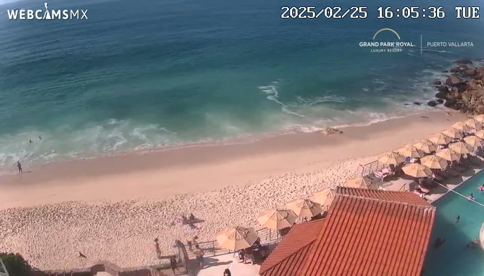 A sandy beach with calm ocean waves, numerous beach umbrellas, and a resort pool is visible on a sunny day.
