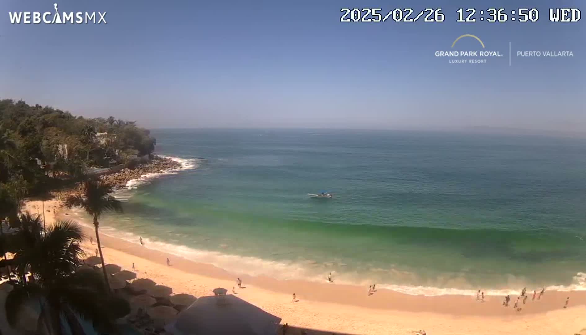 A sandy beach with calm, teal-green ocean waves under a clear, sunny sky is seen from an elevated vantage point, with palm trees and a few people visible.