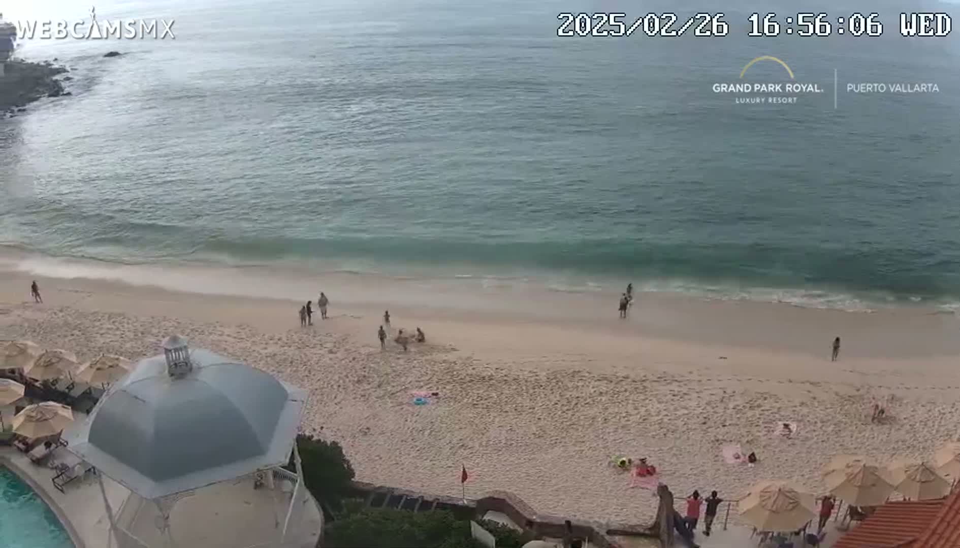An overhead view shows a sandy beach with a few people scattered across it, gentle waves lapping the shore under a mostly cloudy sky.