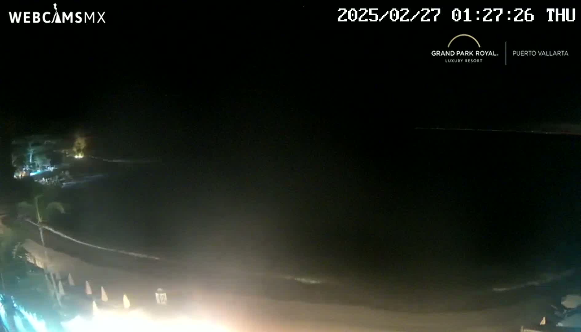 A nighttime, seaside view shows dark water lapping at a sandy shore, with lights illuminating the beachfront area.