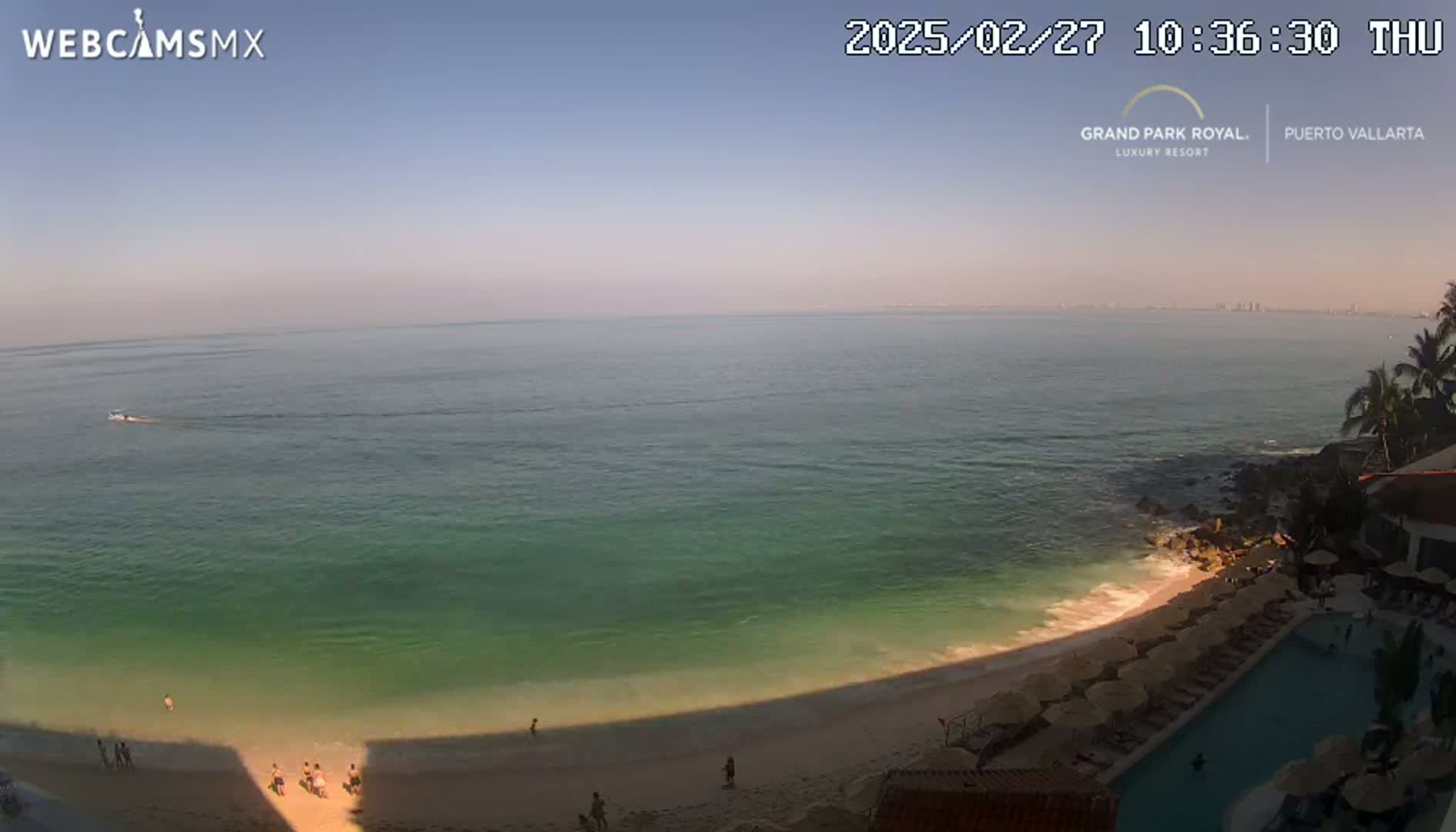A beach with calm, clear turquoise water and a sandy shore, partially shaded by a long shadow, is seen from an elevated vantage point on a sunny day.