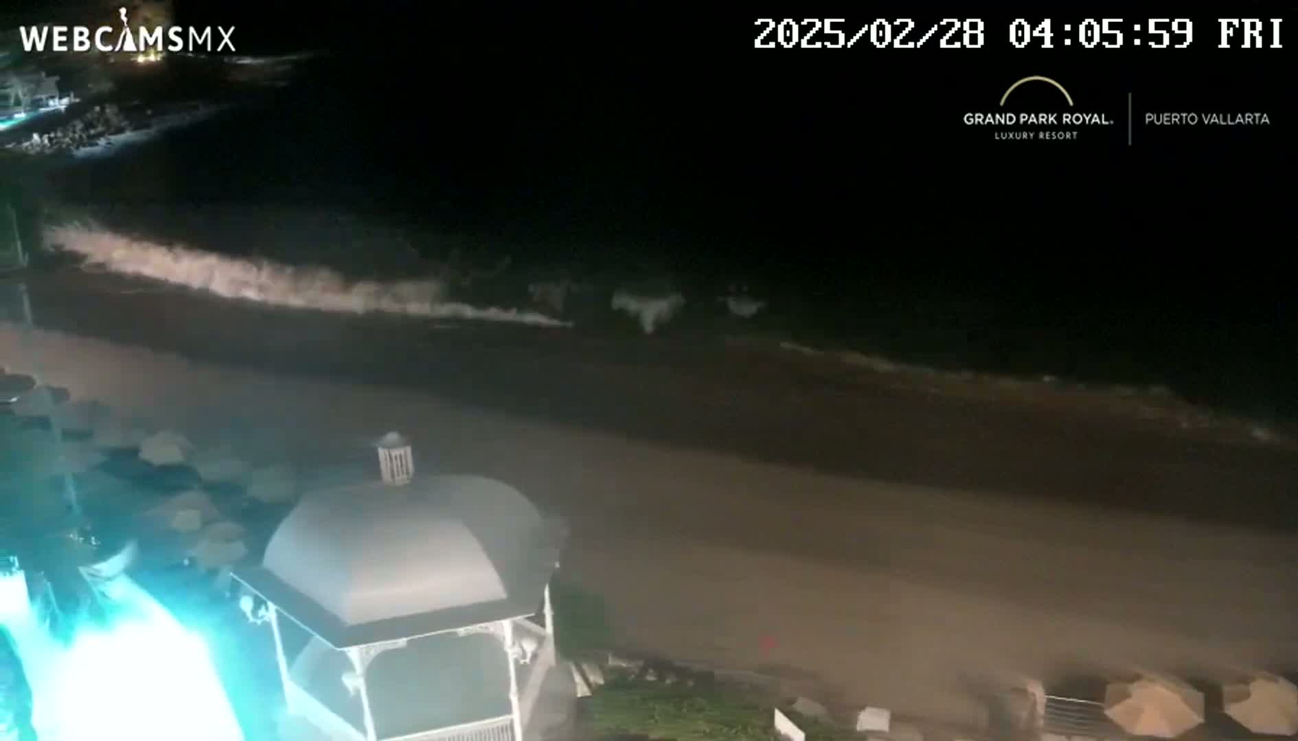 At night, ocean waves roll onto a dark sandy beach near a gazebo, illuminated by a bright teal light.