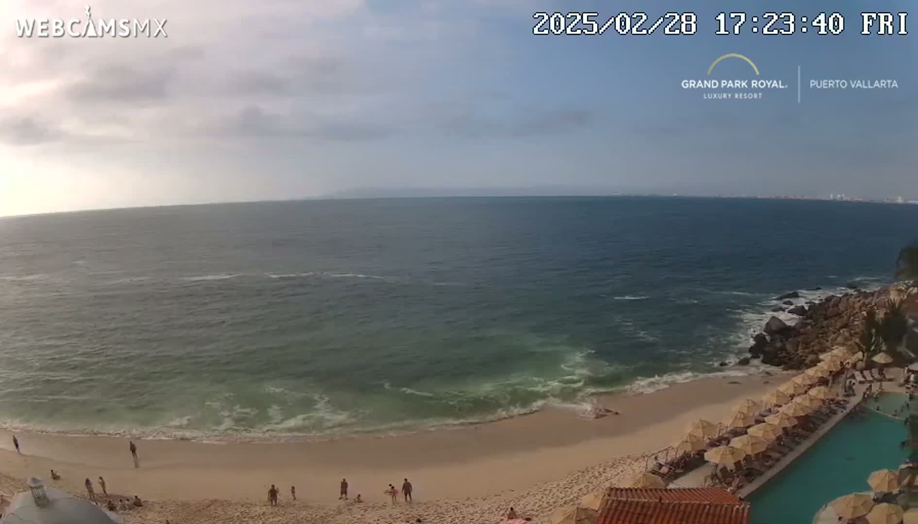 A mostly cloudy day reveals a beach with gently lapping waves, several people, and a resort pool and umbrellas.