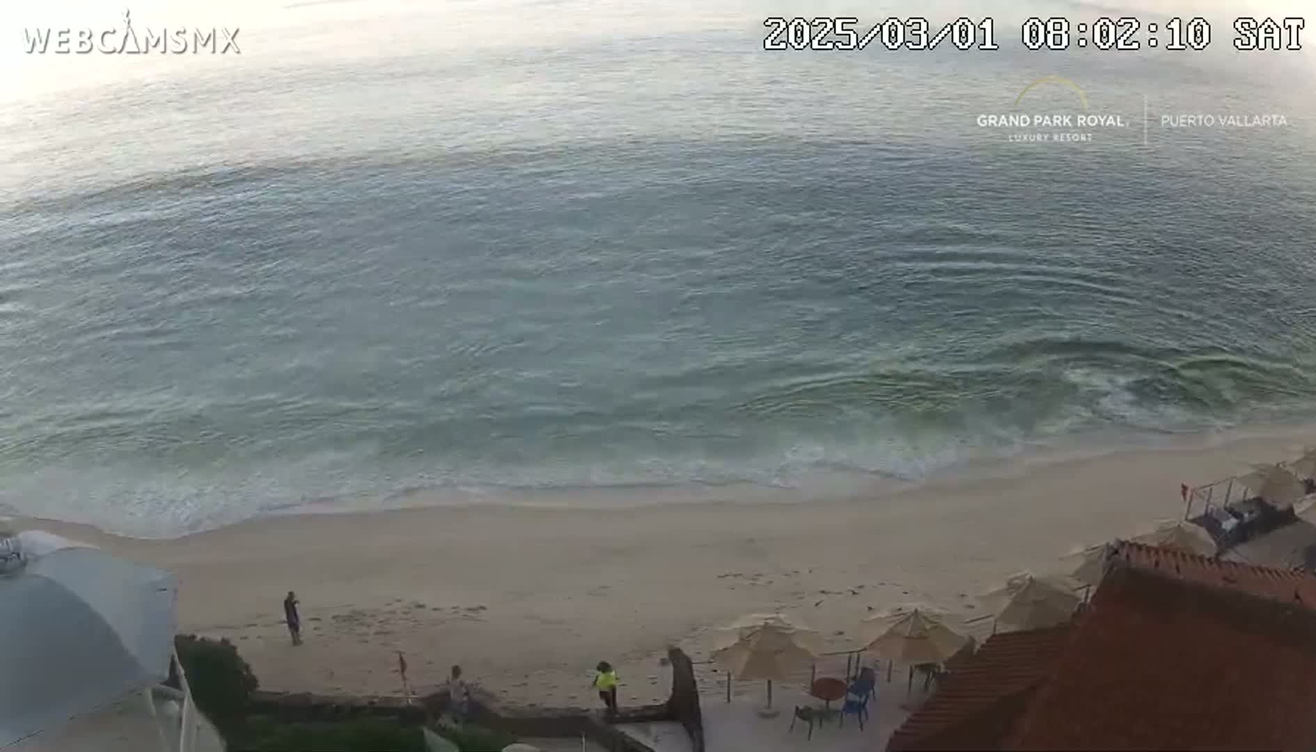 A mostly calm ocean meets a sandy beach where a few people are walking near beach umbrellas and chairs under a partly cloudy sky.