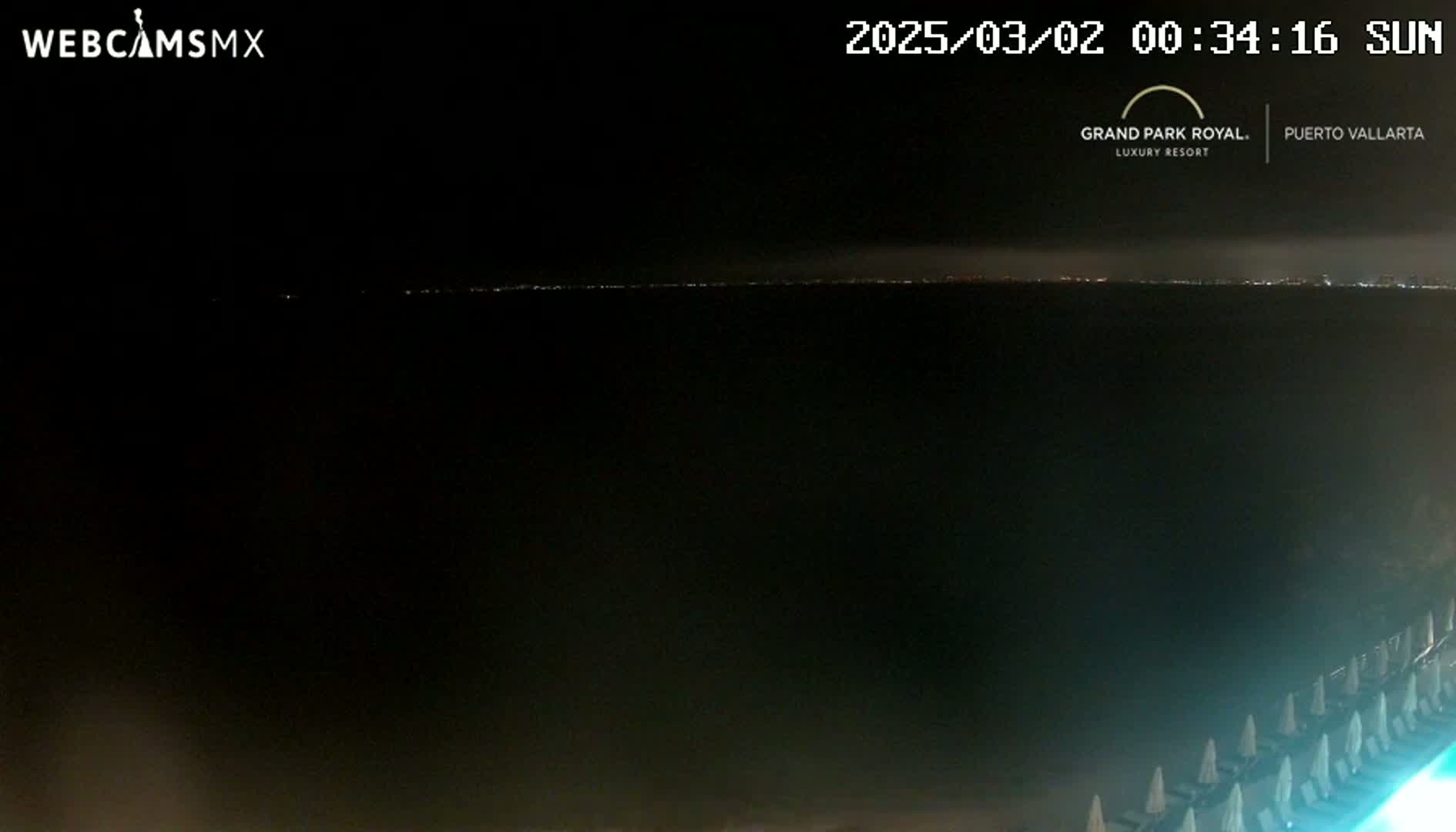 A nighttime view of a dark ocean with a faint line of city lights on the horizon, and beach chairs in the foreground.
