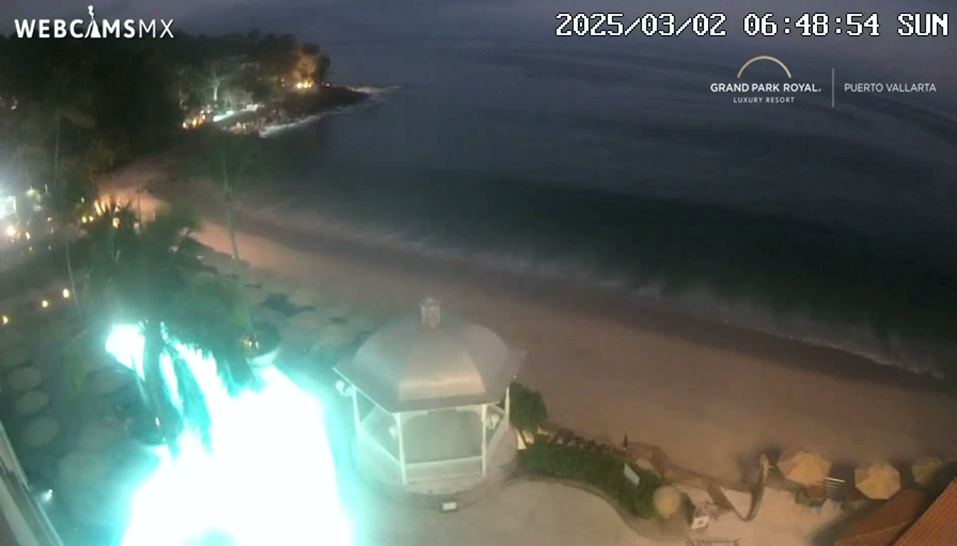 A nighttime view of a beach with gentle waves under a dark sky shows a beachside resort with a gazebo and illuminated landscaping.