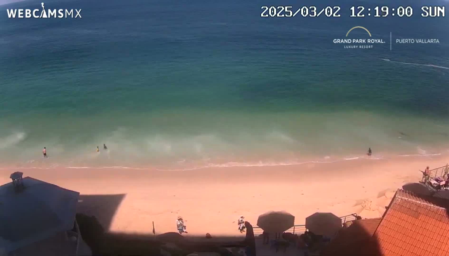 An aerial view shows a sandy beach with a few people on it and in the shallow water, next to a calm, teal ocean under sunny skies.