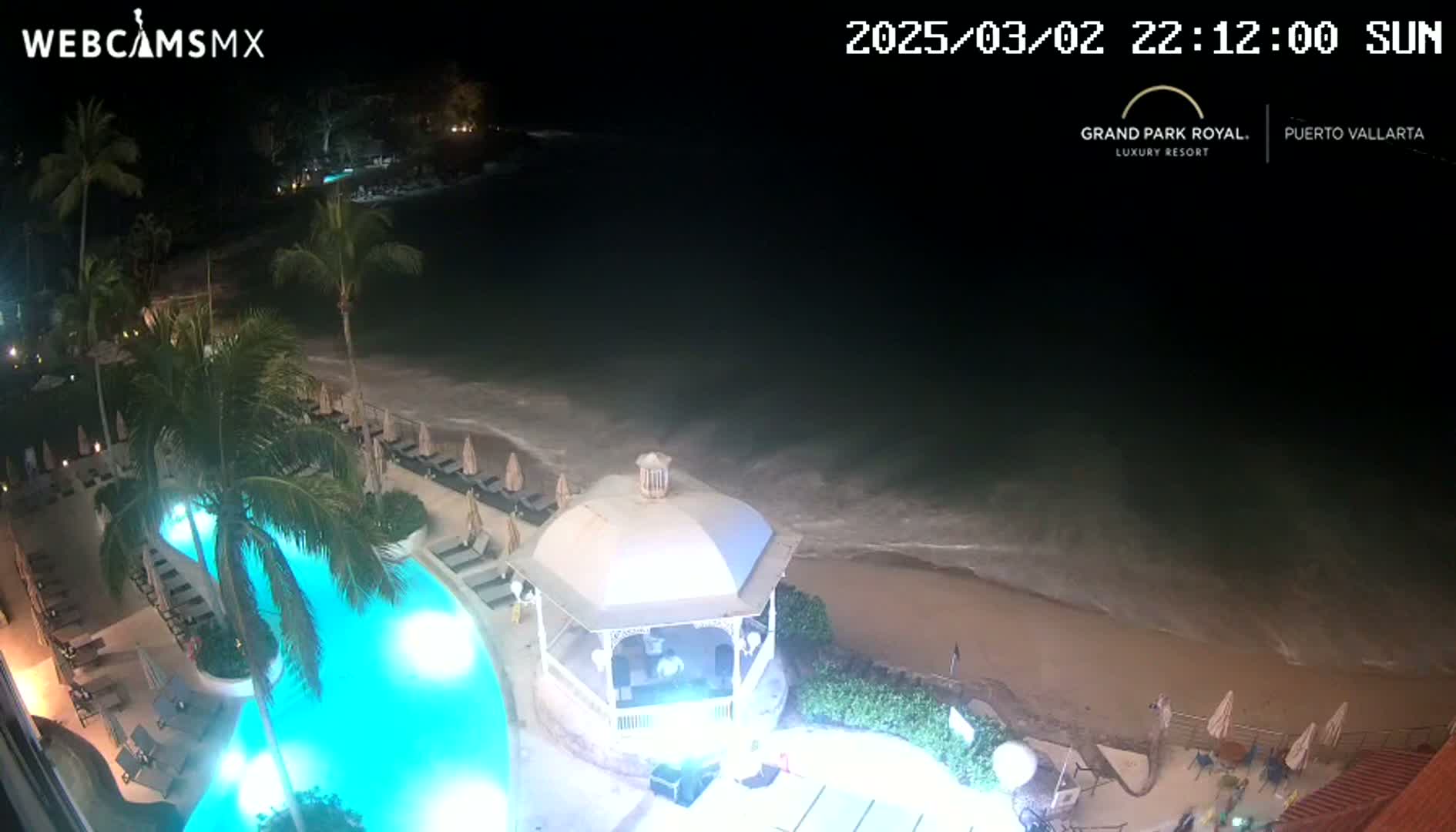 A nighttime aerial view shows a resort pool illuminated in bright blue, next to a beach with gentle waves under a dark sky.