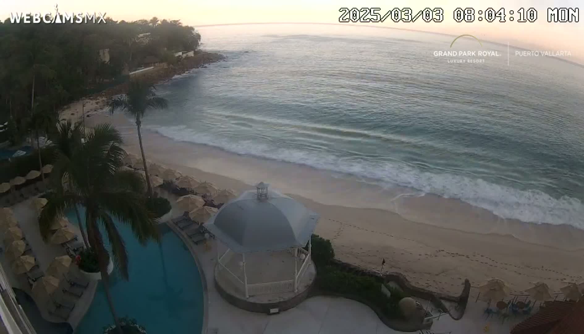 A beach with gentle waves and a resort featuring a swimming pool and a gazebo is seen from an elevated viewpoint at dawn.