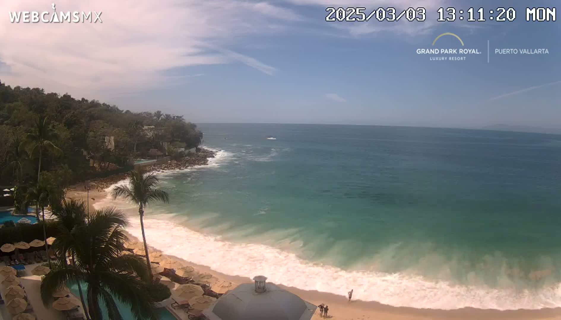 A sandy beach with gentle waves laps the shore under a mostly sunny sky, backed by lush green vegetation and a resort.
