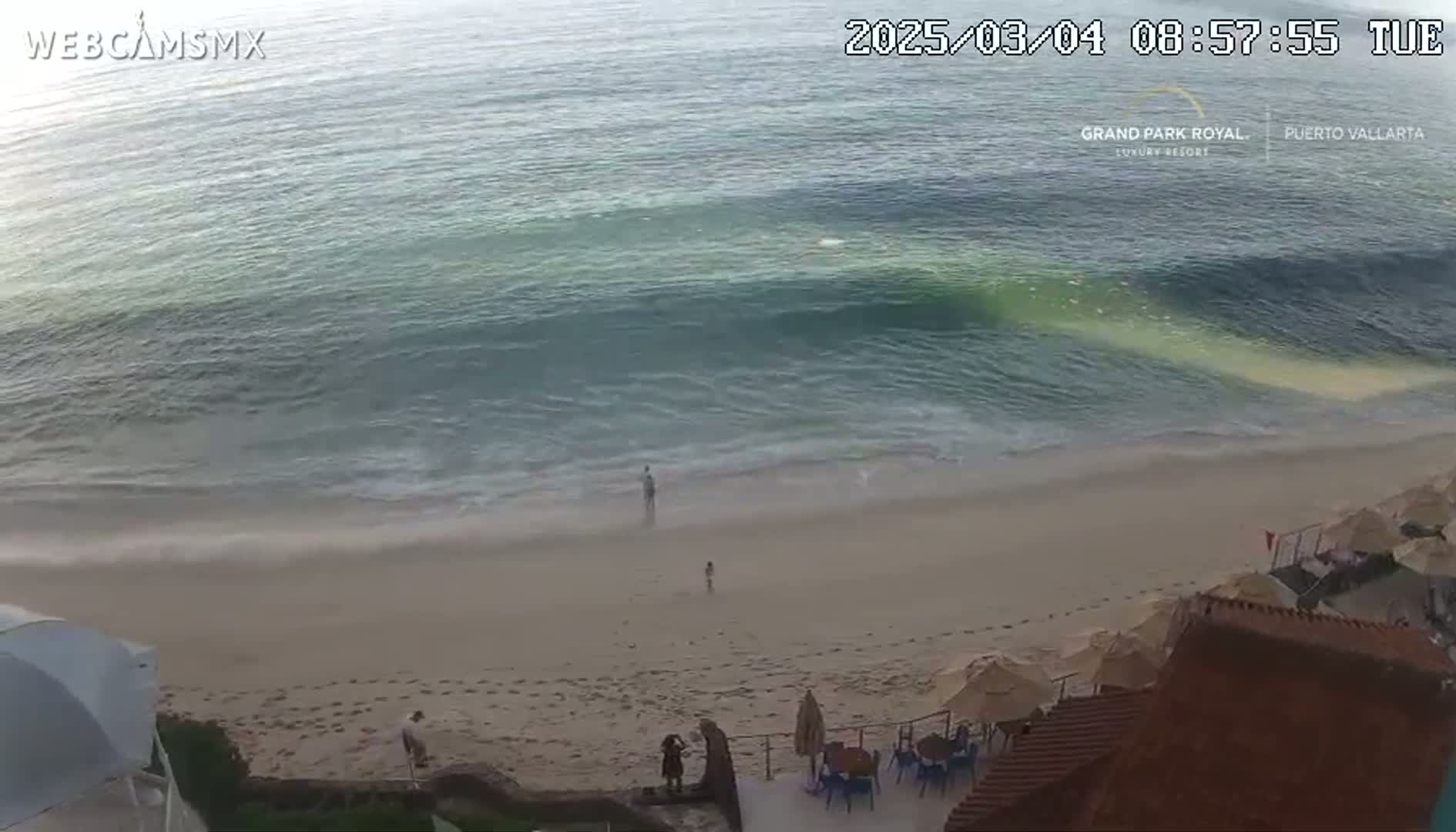 A sandy beach with a few people on it is gently lapped by waves in a calm ocean.