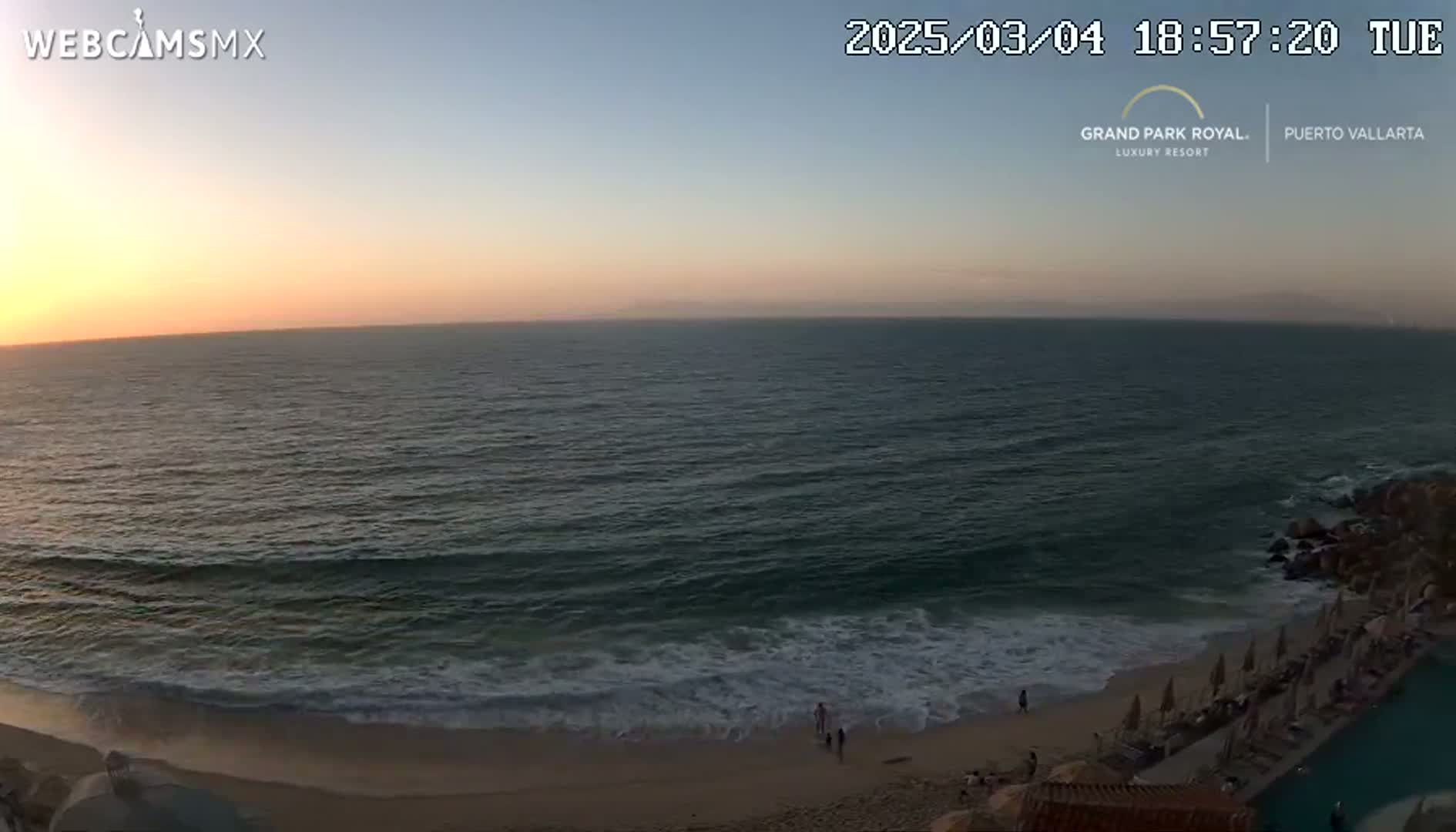 A beach at sunset shows dark-blue ocean waves lapping a sandy shore with a few people on the sand, near a resort.