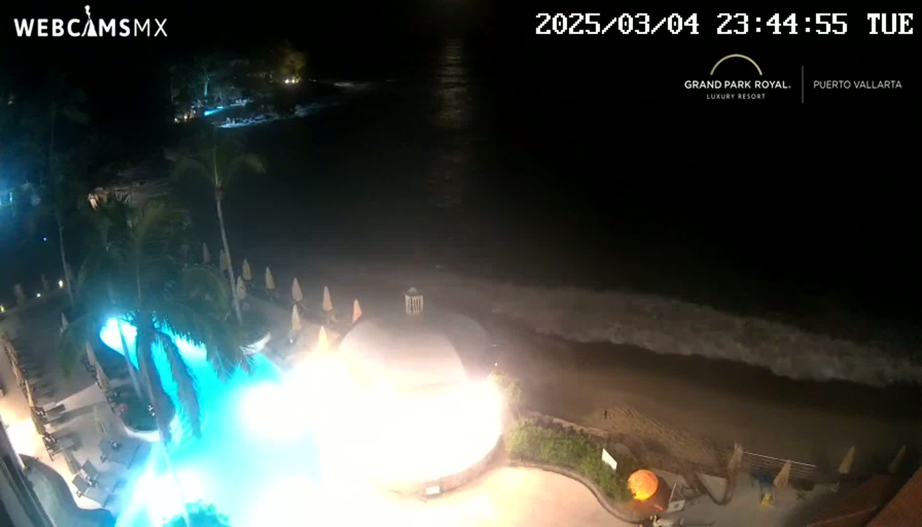 A nighttime aerial view shows a resort pool with bright blue lighting, a beach with gentle waves, and palm trees under a clear, dark sky.