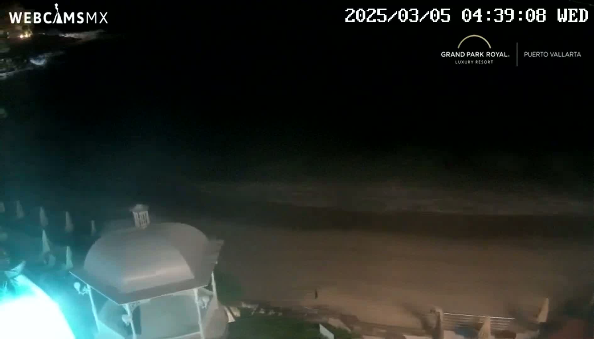 A nighttime view shows a dark beach with gentle waves lapping the shore, next to a building with a domed roof and illuminated by an artificial light source.