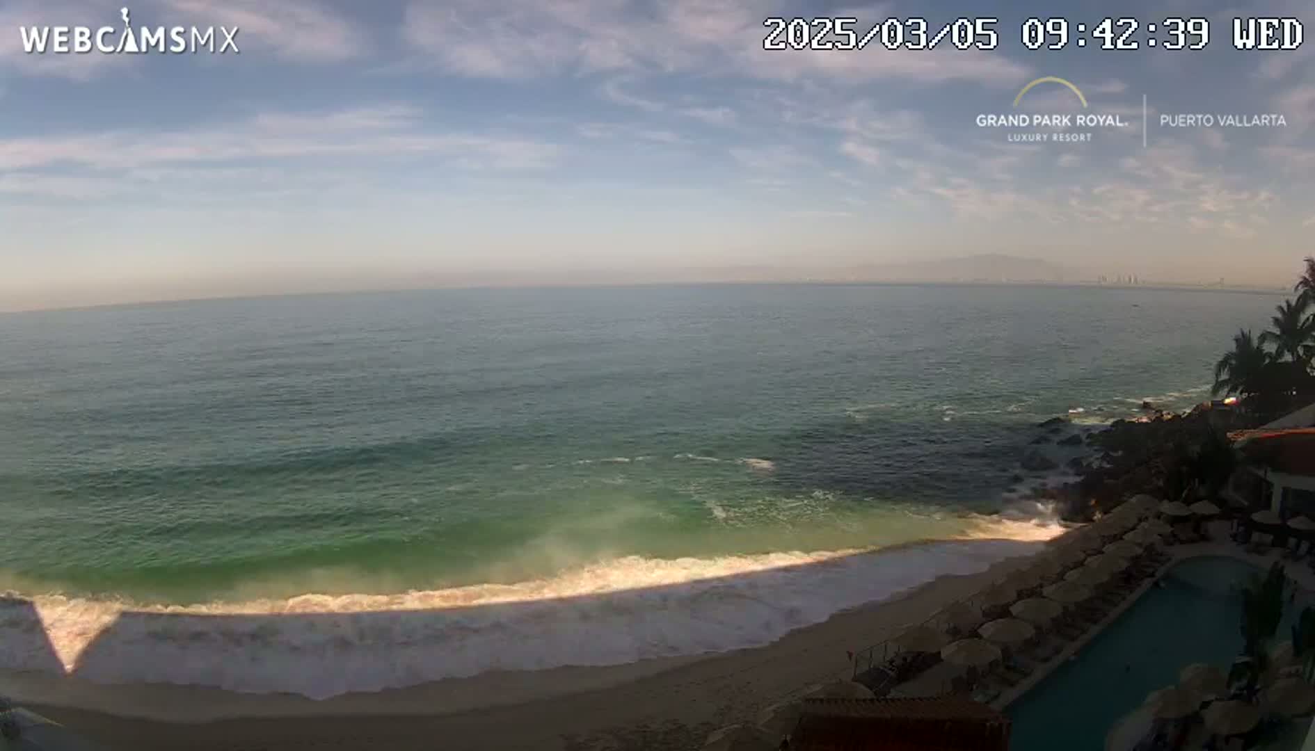 An ocean beach with calm waves, under a partly cloudy sky, is viewed from a resort overlooking the shore.