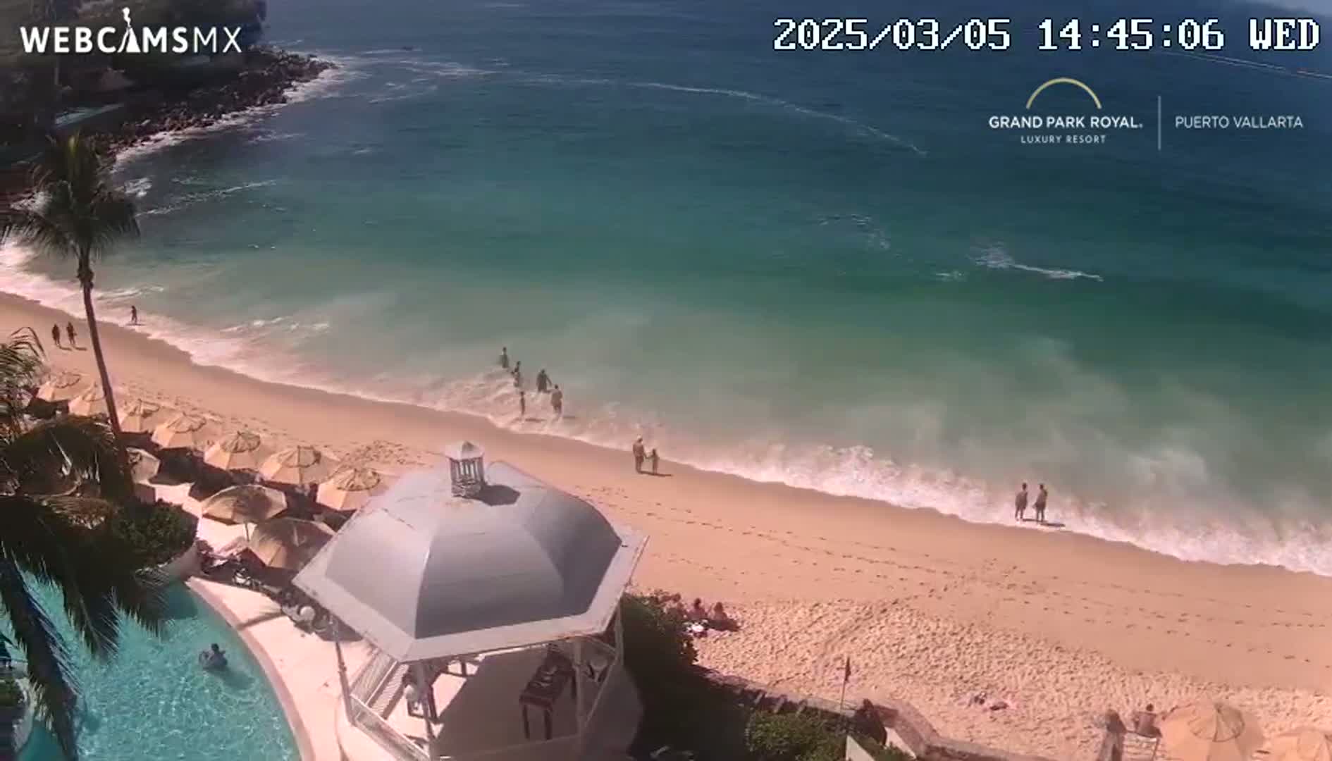 A sandy beach with gentle waves and a few people enjoying the sunny day, next to a resort pool and a gazebo.