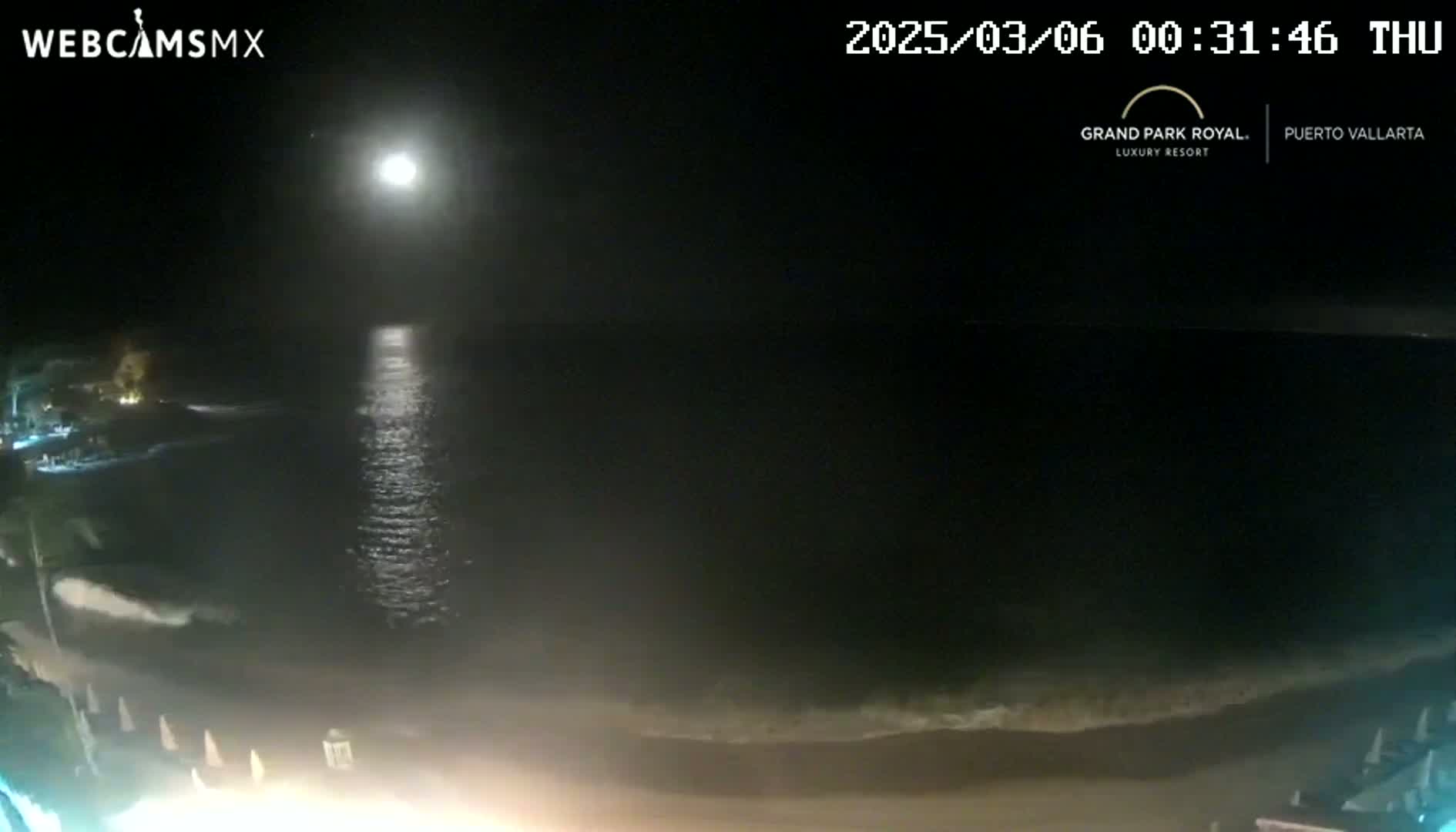 A nighttime beach scene shows a bright moon reflecting on calm ocean water lapping gently at the shore.
