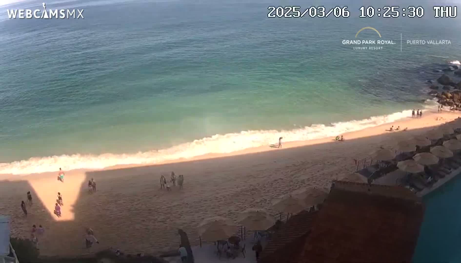 A sandy beach with a few people and umbrellas is next to a calm, teal ocean under sunny skies.